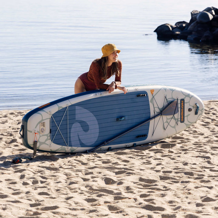 Woman holding a paddleboard on a sandy beach with water in the background.