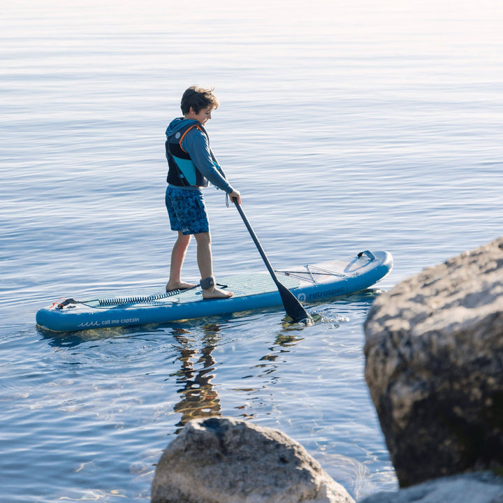 Child paddleboarding on a calm body of water with rocks in the foreground.
