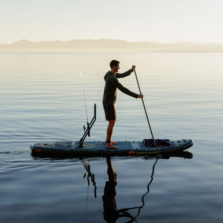 A person stands while paddling their June Inflatable Fishing Paddle Board Kayak Hybrid with the fishing rod holder attached to the board in calm water while the sun it setting.