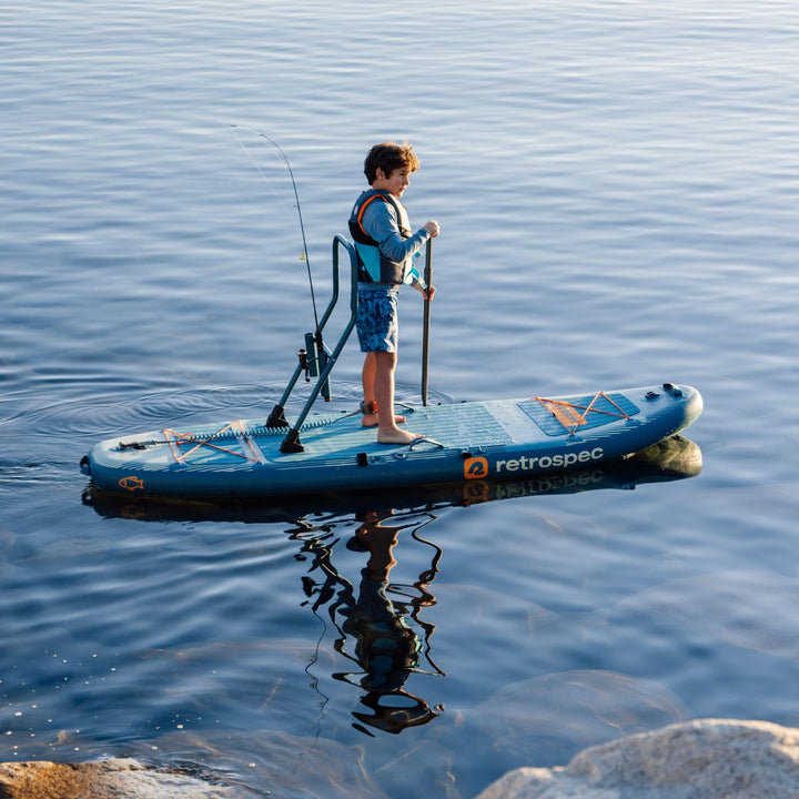 A child paddles while on a June Nano Kids Inflatable Stand Up Paddle Board Kayak Hybrid with 'retrospec' branding on a calm body of water.