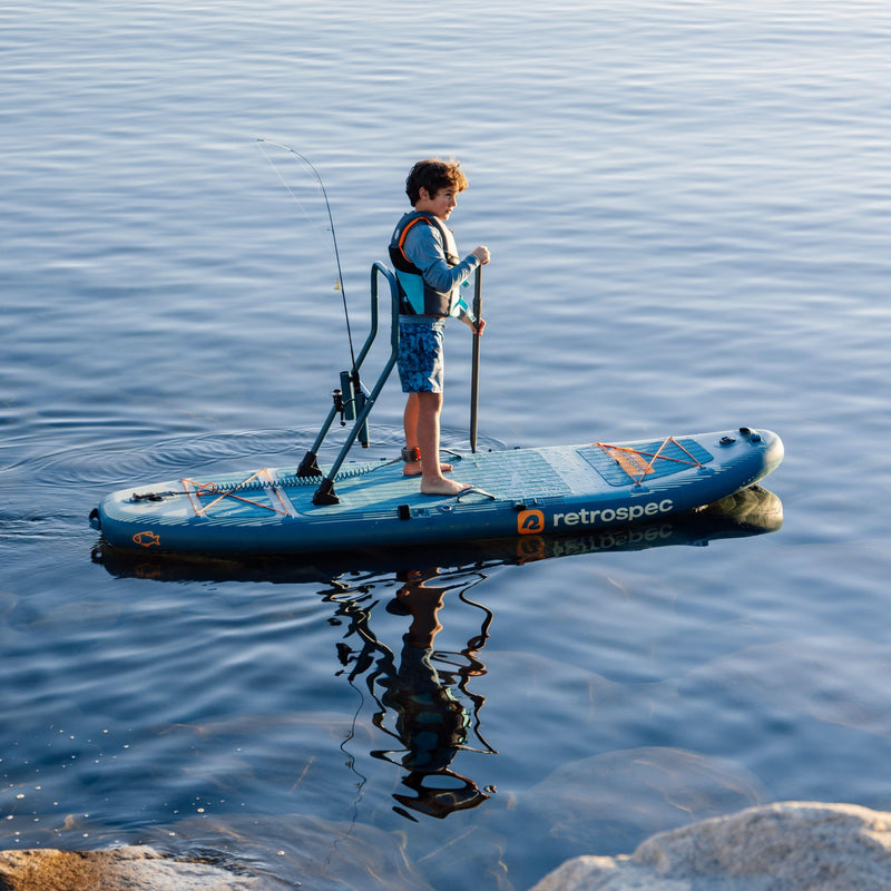 A child paddles while on a June Nano Kids Inflatable Stand Up Paddle Board Kayak Hybrid with 'retrospec' branding on a calm body of water.