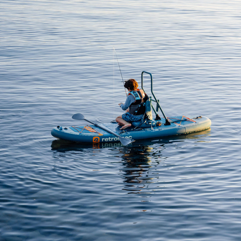 A kid sits on the blue June Nano Kids Inflatable Fishing Paddle Board Kayak Hybrid with fishing equipment on a calm body of water.