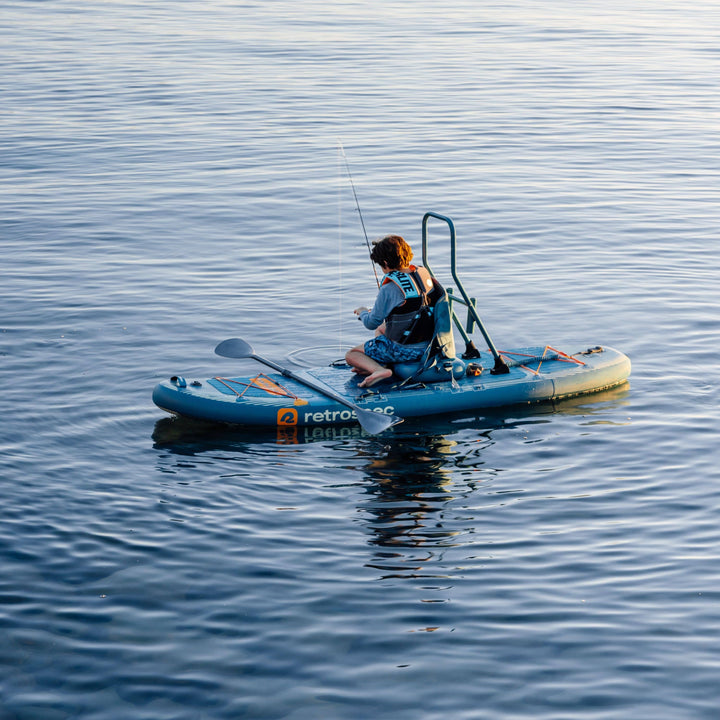 A kid sits on the blue June Nano Kids Inflatable Fishing Paddle Board Kayak Hybrid with fishing equipment on a calm body of water.