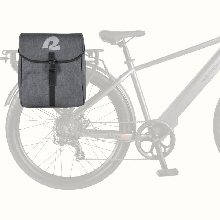 A dark gray bicycle pannier saddle bag with a white reflective retrospec 'R' logo on the front flap, shown mounted on the rear rack of a light gray bicycle against a plain off-white background.