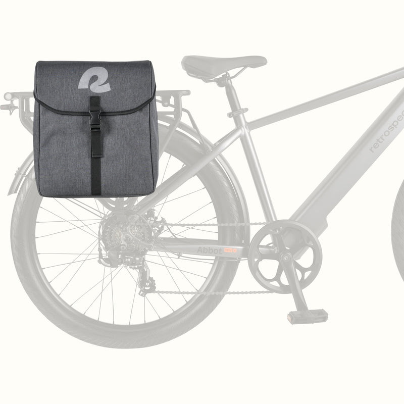 A dark gray bicycle pannier saddle bag with a white reflective retrospec 'R' logo on the front flap, shown mounted on the rear rack of a light gray bicycle against a plain off-white background.