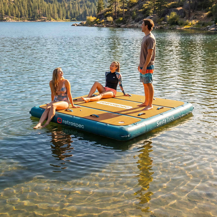 Three friends relaxing on a retrospec Siesta Dock inflatable floating dock on a clear mountain lake surrounded by pine trees.