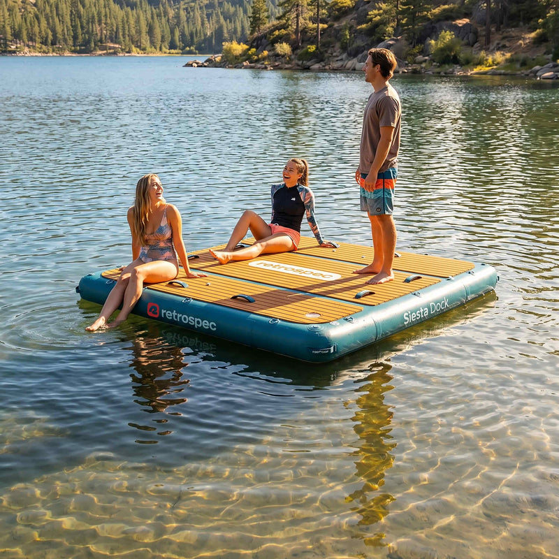 Three friends relaxing on a retrospec Siesta Dock inflatable floating dock on a clear mountain lake surrounded by pine trees.