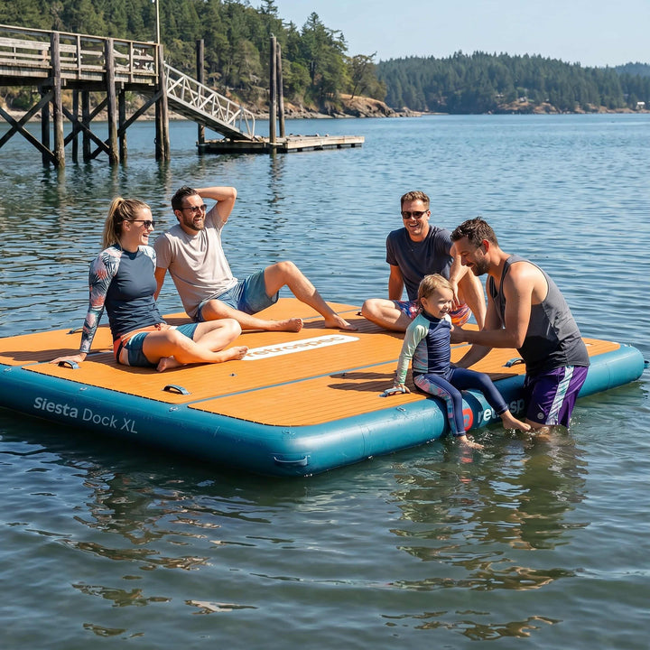 Group of adults and a toddler gathered on a retrospec Siesta Dock XL floating dock for lakes near a wooden pier on a sunny day.