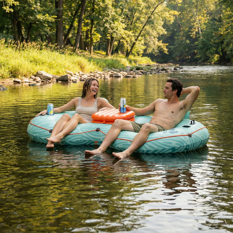 A smiling couple lounging on the retrospec Siesta Float Double on a sunny river, each holding a drink can, with the orange cooler attachment between them and a rocky, tree-lined riverbank in the background.