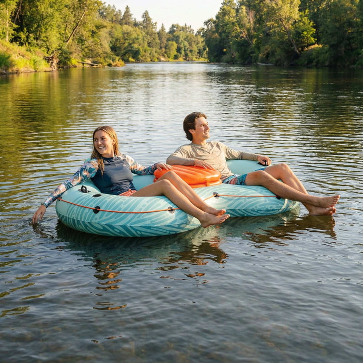 Two people laughing and relaxing on the retrospec Siesta Float Double inflatable river float, drifting down a calm tree-lined river on a sunny day, with an orange cooler attachment between them.