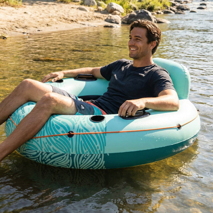A smiling man relaxes on the retrospec Siesta Float inflatable river tube on a calm, sun-dappled river, with a rocky shoreline and green vegetation in the background.