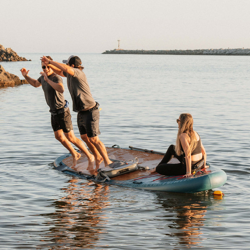 Three friends laughing and enjoying the retrospec Weekender Crew XL inflatable Paddle Board on calm water at golden hour, with two jumping off the wide deck while a third sits at the tail end near a rocky coastline.