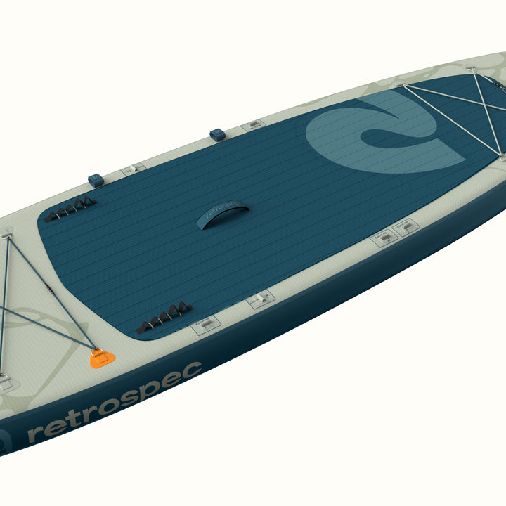 Angled overhead view of the retrospec Weekender Plus Inflatable Stand Up Paddle Board Kayak Hybrid 10'6" in River Ripple Blue, highlighting the full teal textured deck pad, center carry handle, retrospec logo graphic, fin inserts, bungee cargo lacing, and EasyLink accessory mounts along the rail.