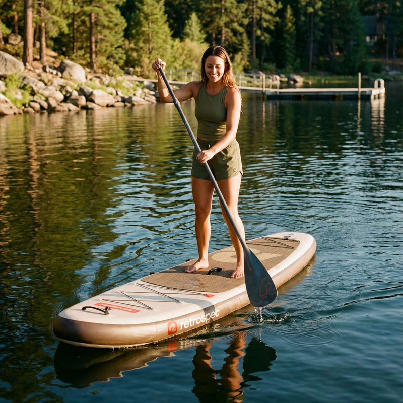 Woman paddleboarding on a calm lake surrounded by trees and rocky shore in warm sunlight.
