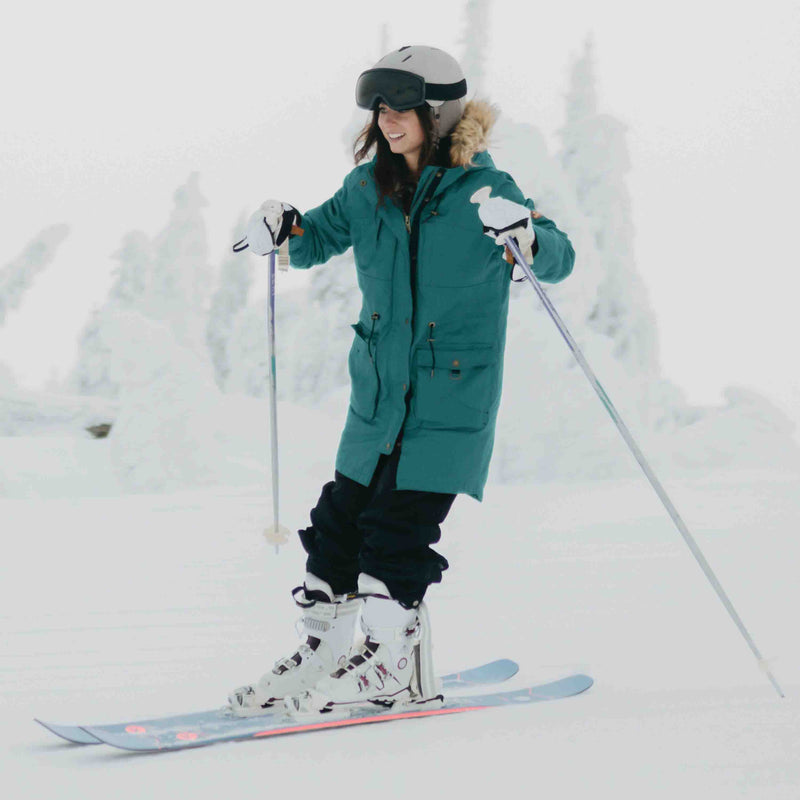 Woman wearing a retrospec Zypher ski helmet and goggles skis down a snowy slope in a teal parka.